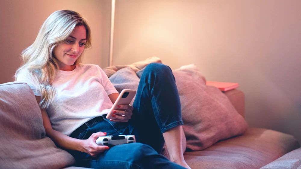 Eine Frau sitzt gemütlich auf dem Sofa und hält in der einen Hand einen Gamecontroller, in der anderen ein Smartphone